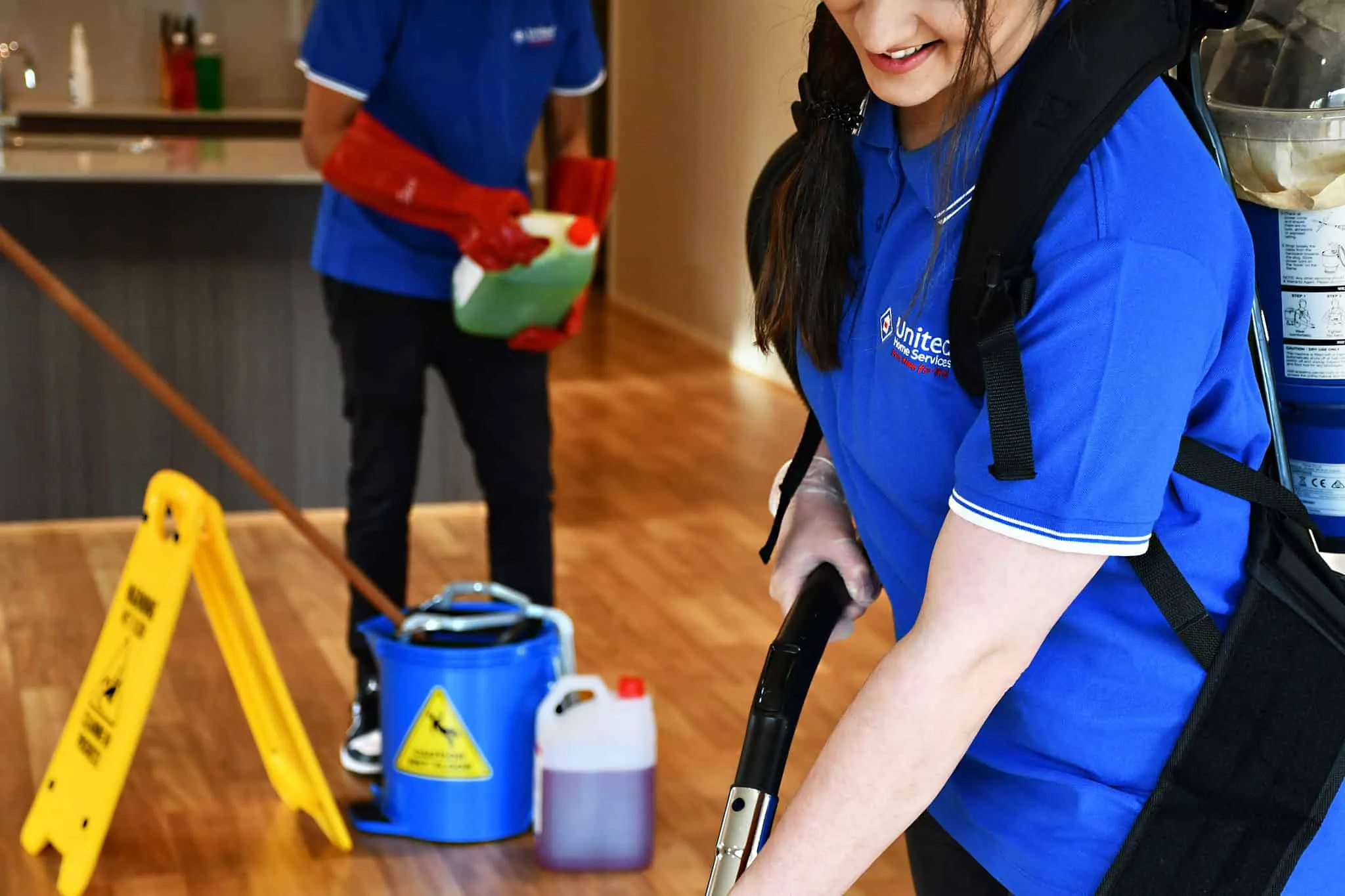 Cleaning team members in blue uniforms using professional equipment, including a vacuum cleaner, cleaning supplies, and caution signs, in a home service setting.