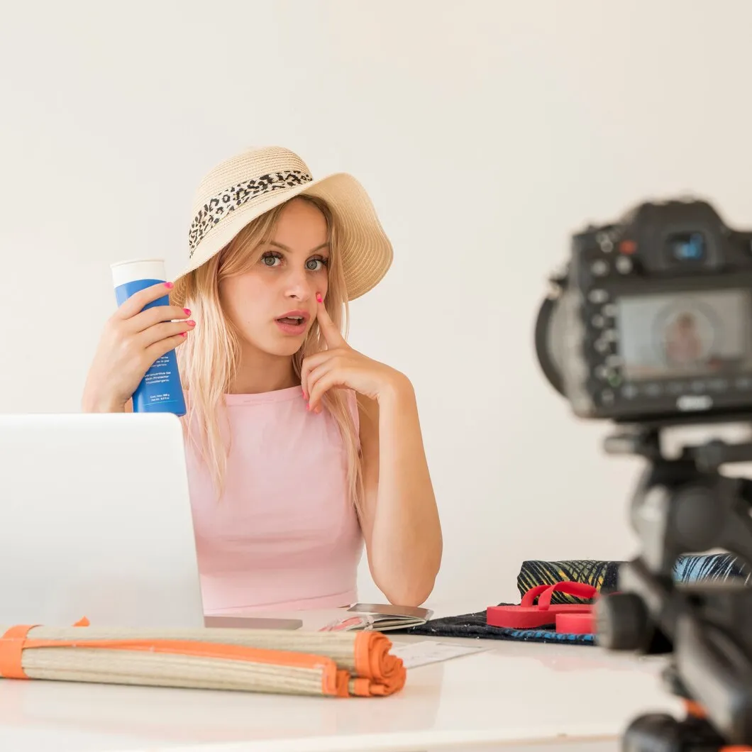 Blonde influencer in a straw hat recording a video while holding a blue product, with a laptop and camera on the table, representing video production and content creation.