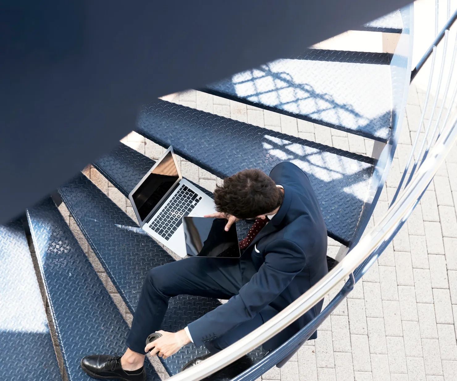 Business professional in a suit working on a laptop and tablet while seated on a spiral staircase, symbolizing digital solutions and modern work environments.