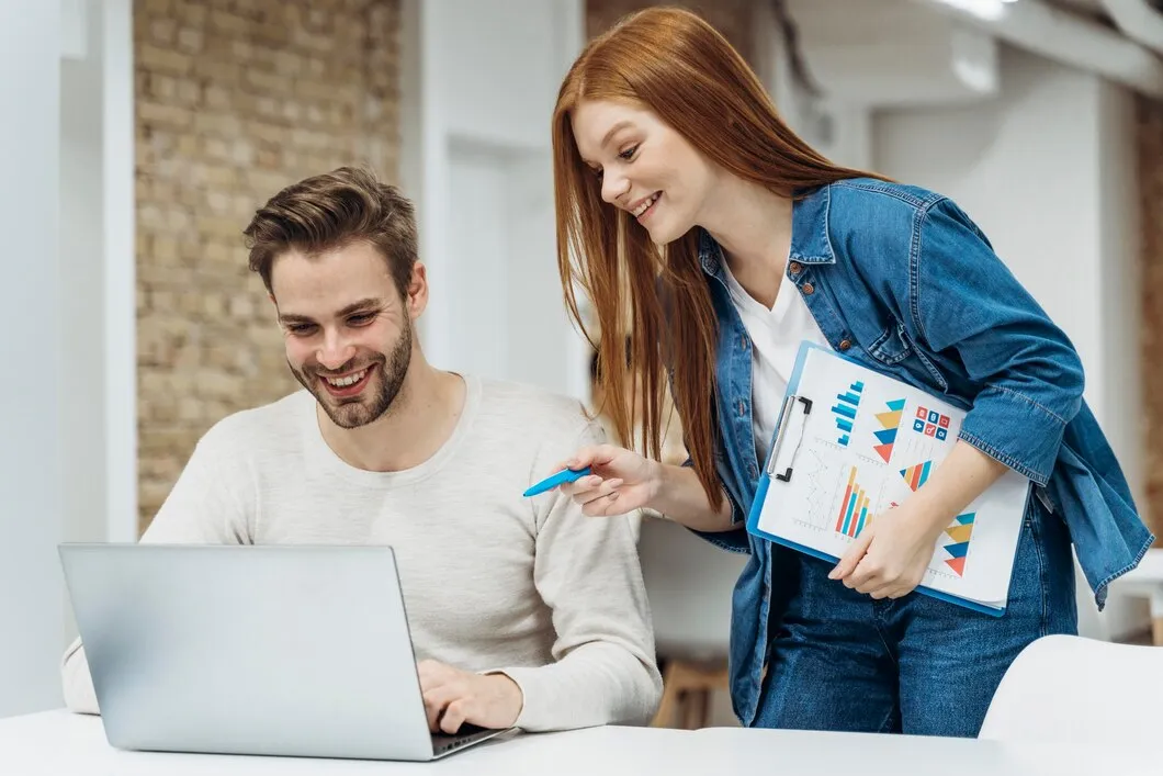 Man and woman discussing business strategies while reviewing data on a laptop, with charts and graphs on a clipboard, in a modern office setting.