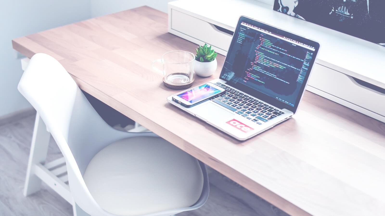 Laptop displaying code on a wooden desk with a smartphone, glass cup, and potted plant, representing digital design and creativity for web development.