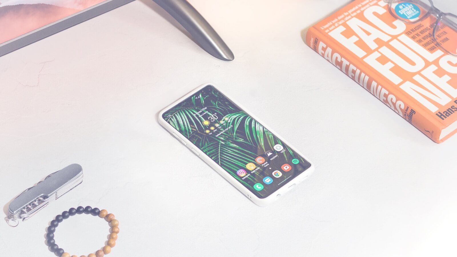 Smartphone displaying a tropical wallpaper alongside a book titled "Factfulness," a multi-tool, and a wooden bracelet, emphasizing modern design and digital connectivity.