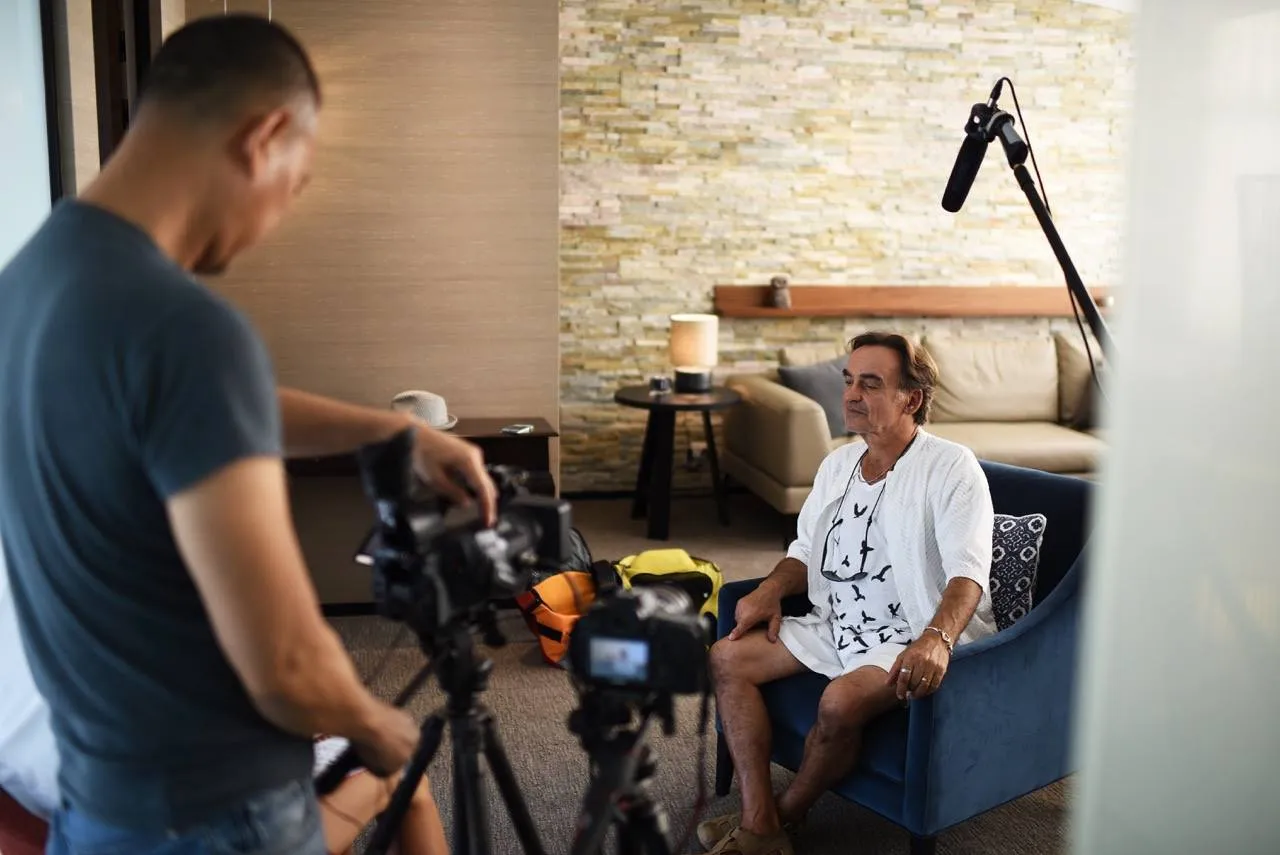 Video production setup with a man seated in a blue chair, being filmed by a crew member holding a camera, and a microphone positioned overhead, in a well-lit indoor setting.