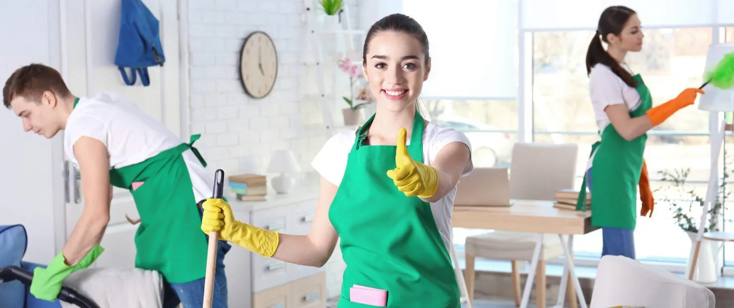 Three smiling cleaning professionals in green aprons and yellow gloves, actively engaged in cleaning tasks within a bright, modern home environment, emphasizing home service expertise and customer satisfaction.