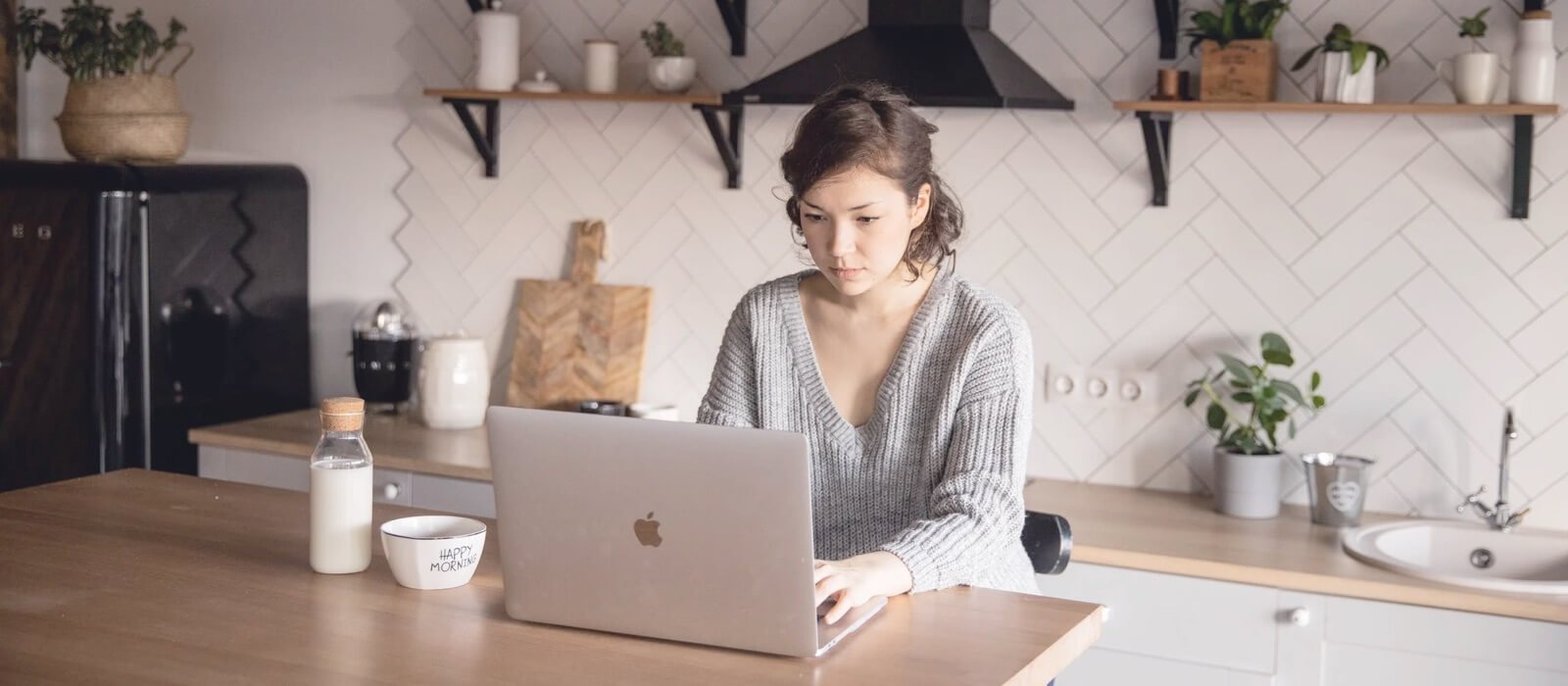 Woman working on a laptop in a modern kitchen, surrounded by plants and kitchenware, illustrating a creative workspace for digital services and web design.