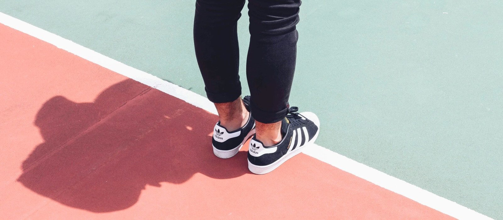 Person wearing black jeans and Adidas sneakers standing on a tennis court with red and green surfaces, showcasing sporty style and active lifestyle.