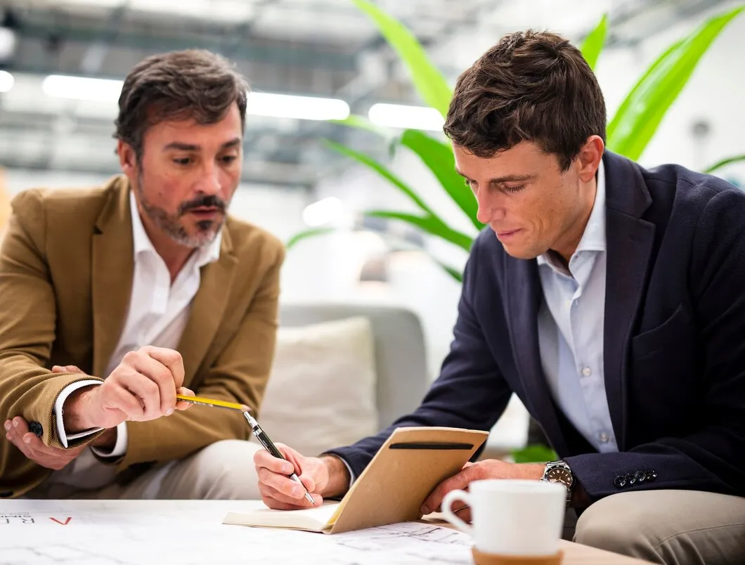 Male coworkers discussing strategies for review and reputation management, one taking notes on a notepad, with a coffee cup and plant in the background.